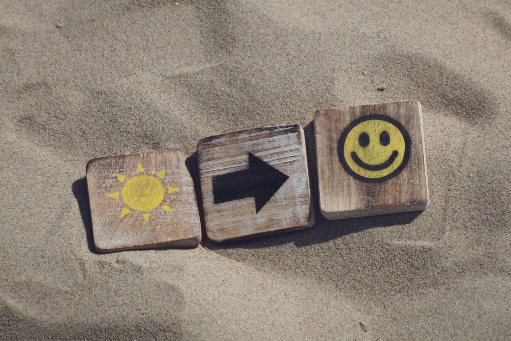 Wooden blocks in the sand showing a sun, an arrow, and a smiley face, symbolizing improved mood and well-being through vitamin D— a visual concept supporting hormone balance and midlife wellness.