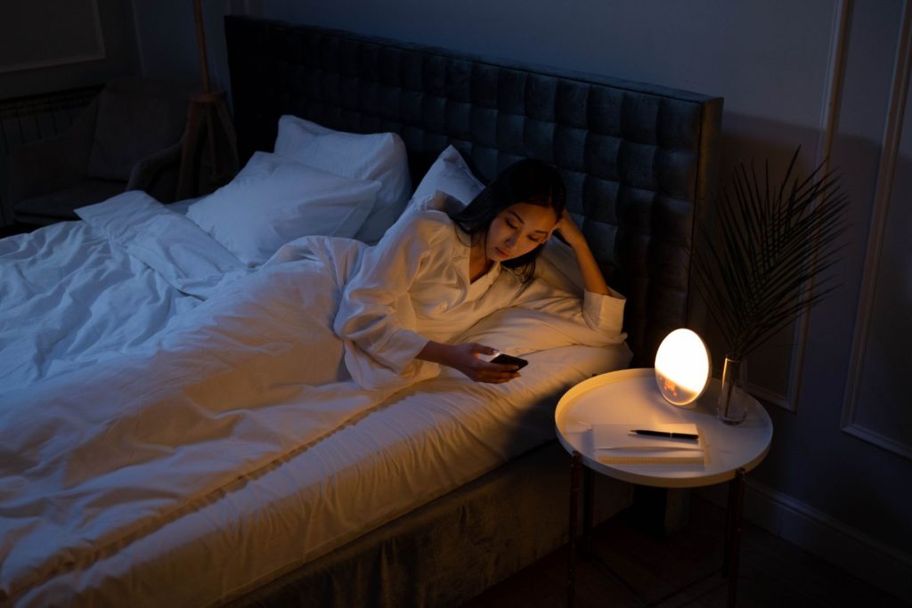Woman lying in bed at night looking at her phone with a bedside lamp, illustrating sleep disruption and how stress affects hormone balance after 40.
