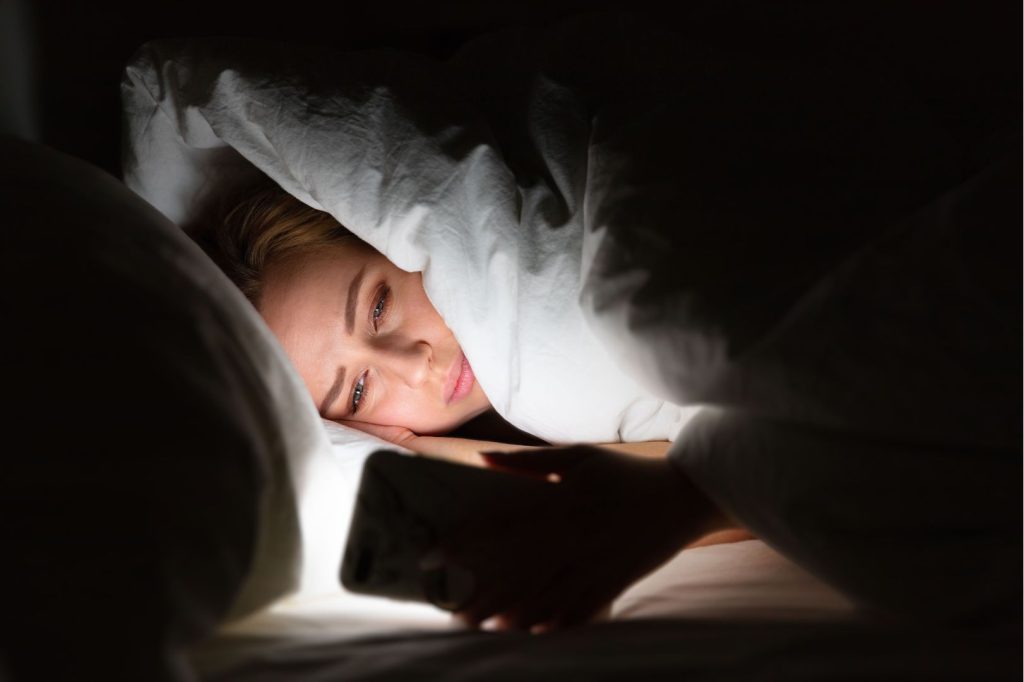 Woman lying awake in bed using her phone at night, illustrating how late-night screen use can disrupt sleep and interfere with a calming evening routine