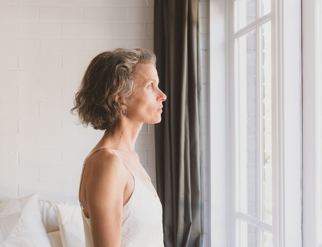 Woman over 50 standing by a window in a calm, reflective moment during postmenopause