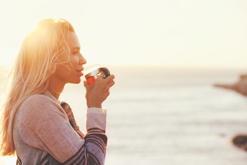 Woman drinking tea at sunrise by the ocean, reflecting on resetting hormones after birth control
