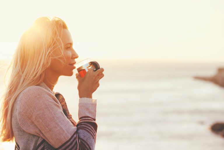 Woman drinking tea at sunrise by the ocean, reflecting on resetting hormones after birth control